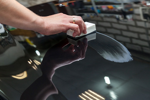 The process of applying a nano-ceramic coating on the car's hood by a male worker with a sponge and special chemical composition to protect the paint on the body from scratches, chips and damage. Concept image of “Is Ceramic Coating Good For My Car?” | Harv’s Auto Body Repair in Iowa City, IA.